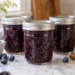 Three sealed canning jars of blueberry-spice jam with fresh blueberries and cinnamon