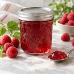 Sealed jar of clear ruby raspberry jelly with fresh raspberries and a spoonful of jelly